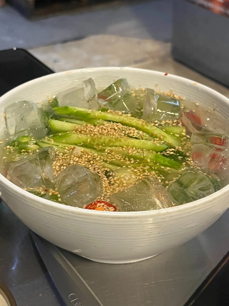 A refreshing bowl of Oiji Naengguksu, a Korean cold noodle soup made with pickled cucumbers, floating ice cubes, and sesame seeds. The tangy and icy broth cuts through the richness of grilled meats, making it a perfect palate cleanser.