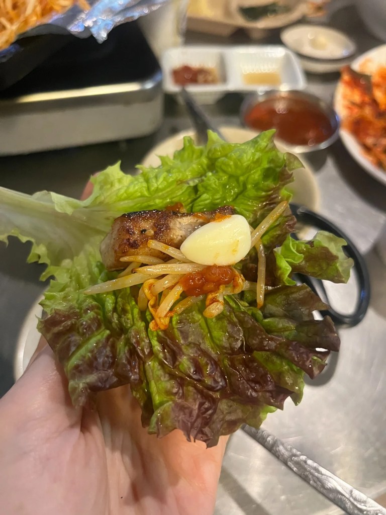 A perfectly assembled Korean BBQ ssam (wrap) featuring a piece of grilled pork, seasoned bean sprouts, fresh garlic, and ssamjang (spicy dipping sauce) wrapped in a crisp lettuce leaf. This traditional Korean way of eating BBQ enhances the flavors and textures in every bite.
