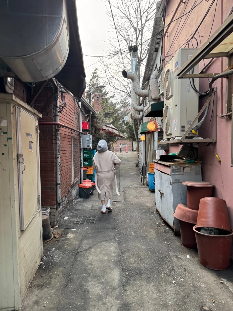 A narrow alleyway beside Sangroksu in Yongsan, Seoul, showcasing the authentic backstreets of old Korean neighborhoods. A local resident walks through, adding to the nostalgic and everyday charm of the area.