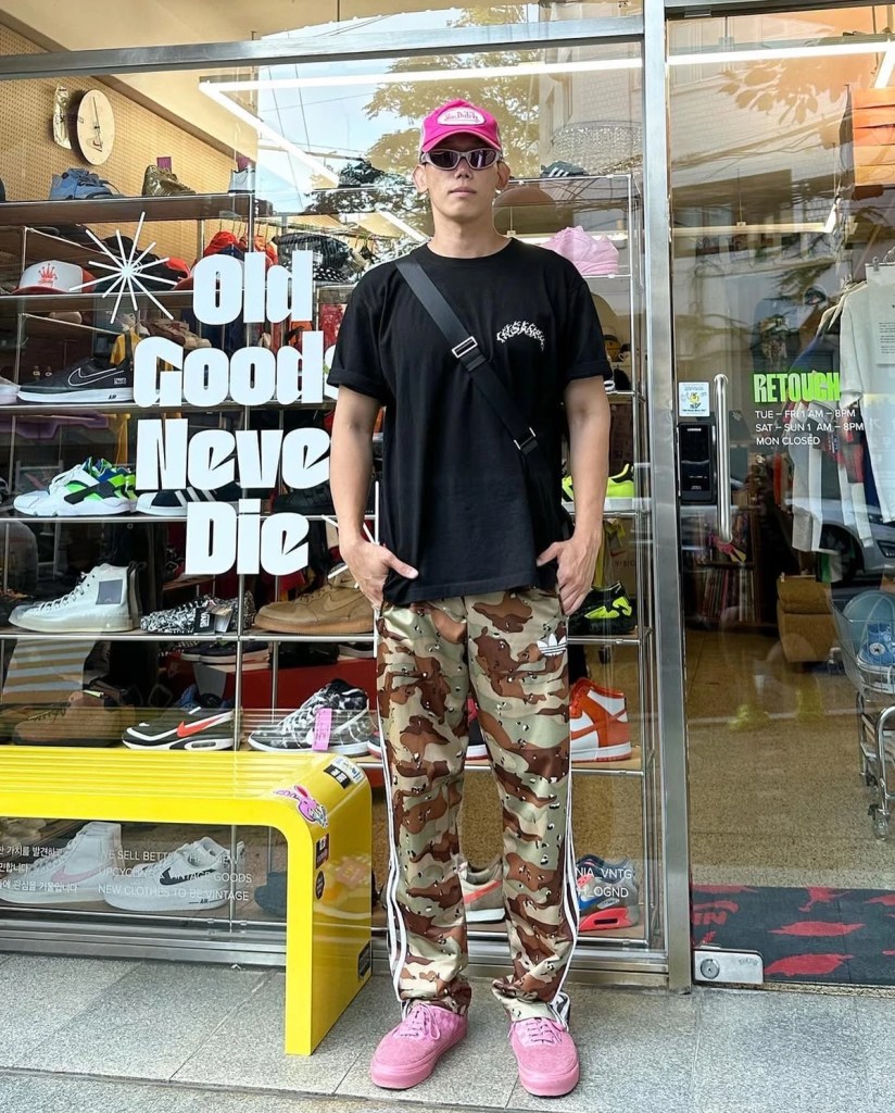 A streetwear enthusiast in a black oversized T-shirt, camo pants, and pink sneakers, standing in front of Retouch Seoul with 'Old Goods Never Die' signage