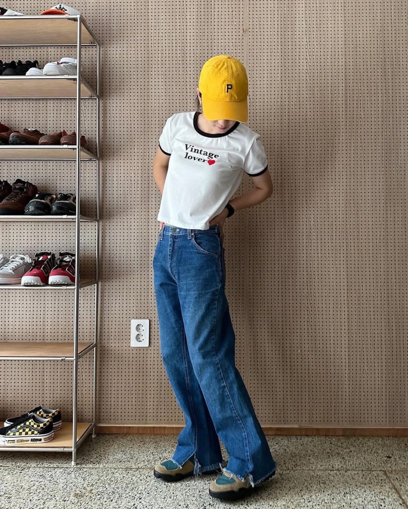 A stylish woman wearing wide-leg jeans, a vintage graphic tee, and a yellow Retouch cap, posing next to a sneaker display at Retouch Seoul