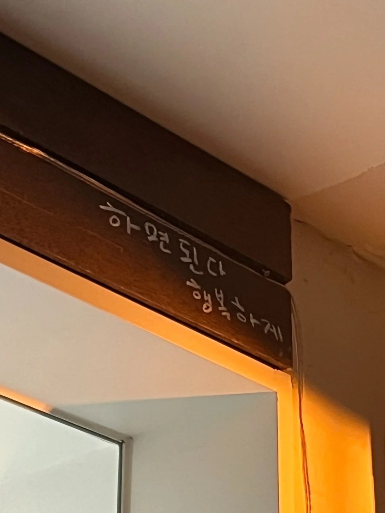 A handwritten message on a wooden beam inside Guden Guden Cafe in Yongsan, Seoul, reading "You can do it, be happy" in Korean. A meaningful motto reflecting the cafe owner's philosophy.